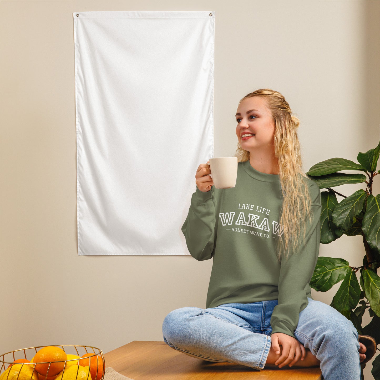 Smiling woman in olive green crewneck sweatshirt with lake life text, sitting on table with coffee cup