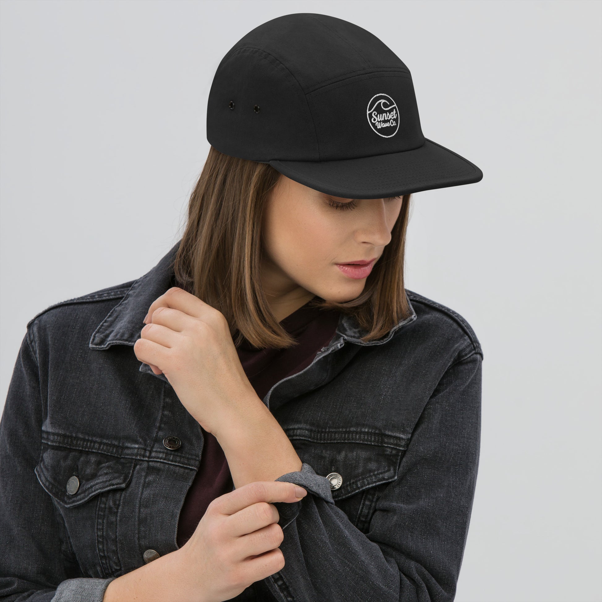 Woman wearing black 5-panel cap with white Sunset Wave Co logo and black denim jacket