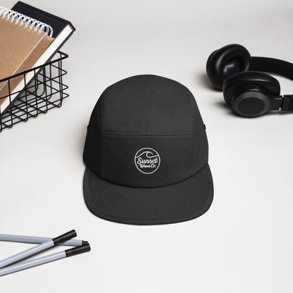 Black 5-panel cap with white Sunset Wave Co. logo, placed on white desk near headphones, pens, and notebooks