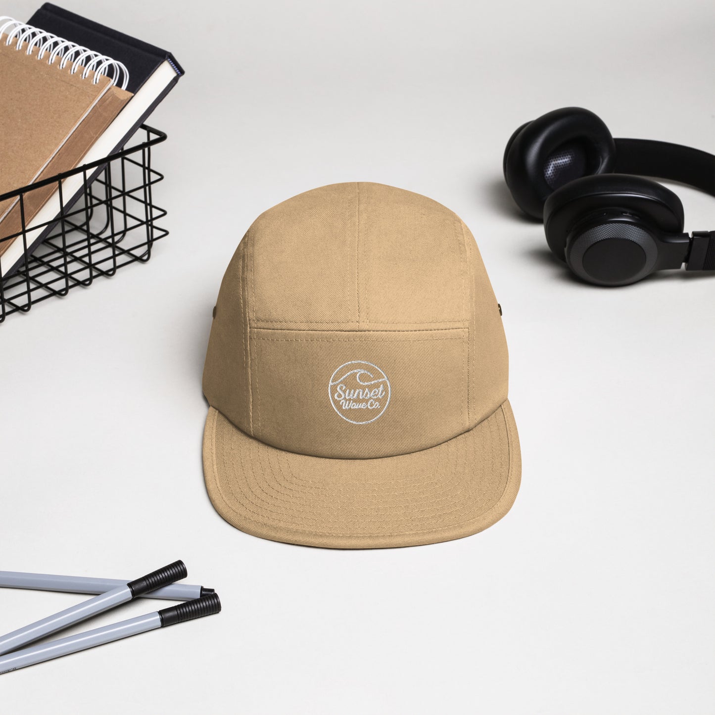 Khaki 5-panel cap with white Sunset Wave Co. logo, black headphones, notebooks, and pens on white desk