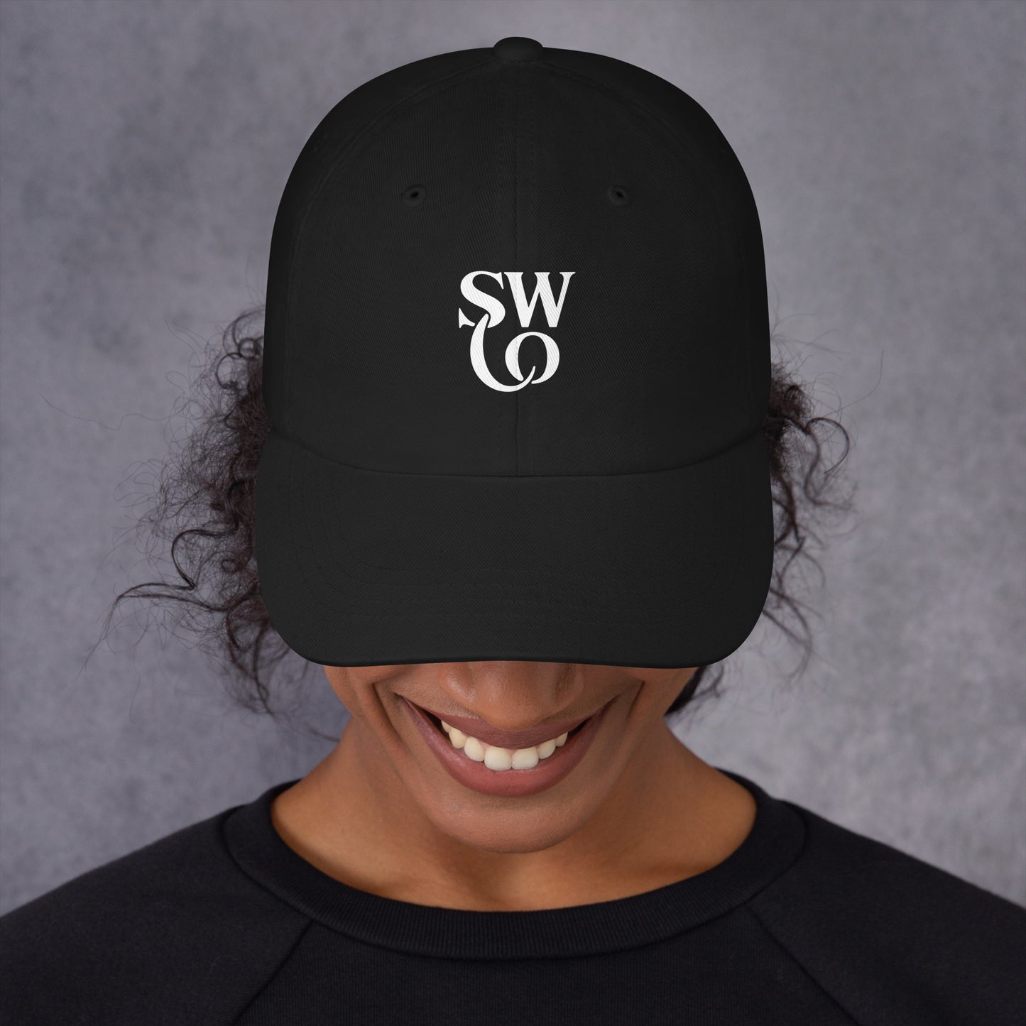Smiling person wearing black dad hat with white SWO logo on front, gray background