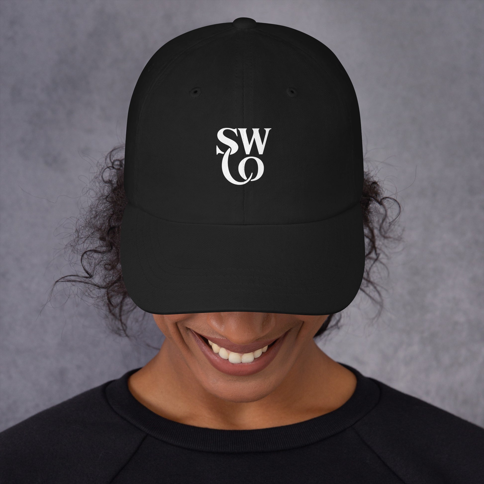 Smiling person wearing black dad hat with white SWO logo on front, gray background