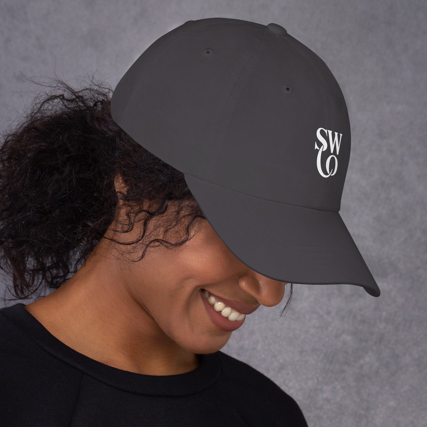 Smiling woman wearing dark grey dad hat with white SWO logo on right side against grey background