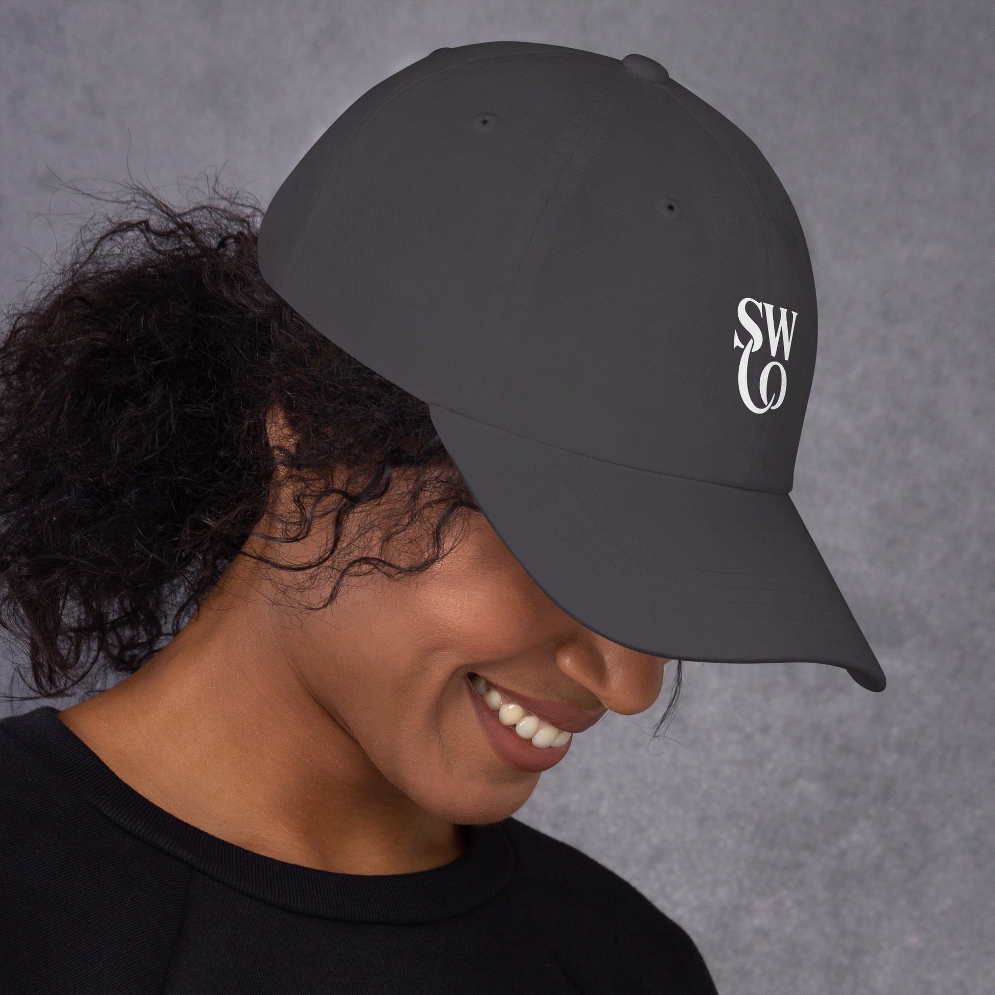 Smiling woman wearing dark grey dad hat with white SWO logo on right side against grey background