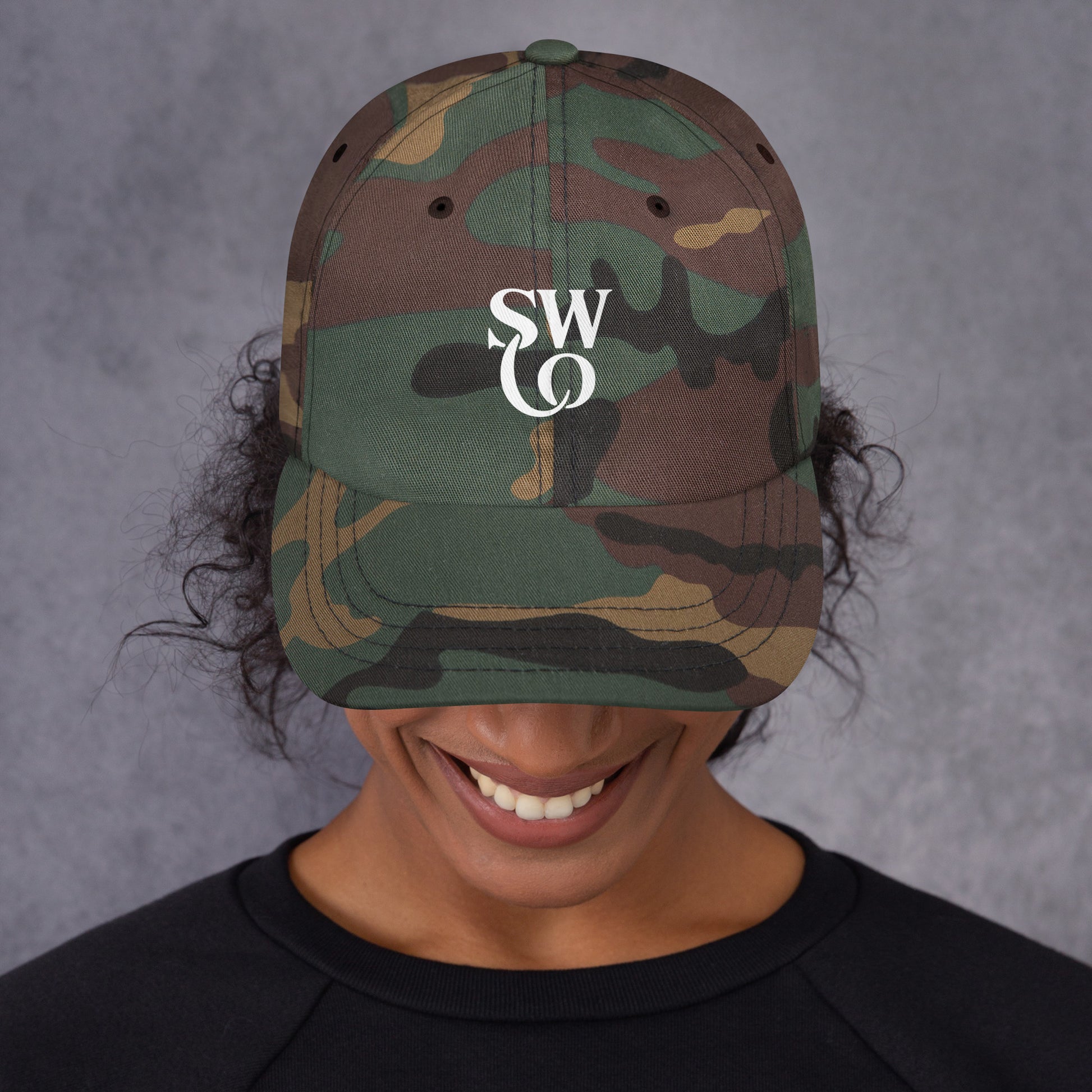 Smiling person wearing green camo classic dad hat with white SWO logo and black shirt