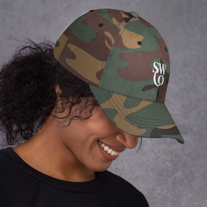 Smiling woman wearing green camo classic dad hat with SWO logo on gray background
