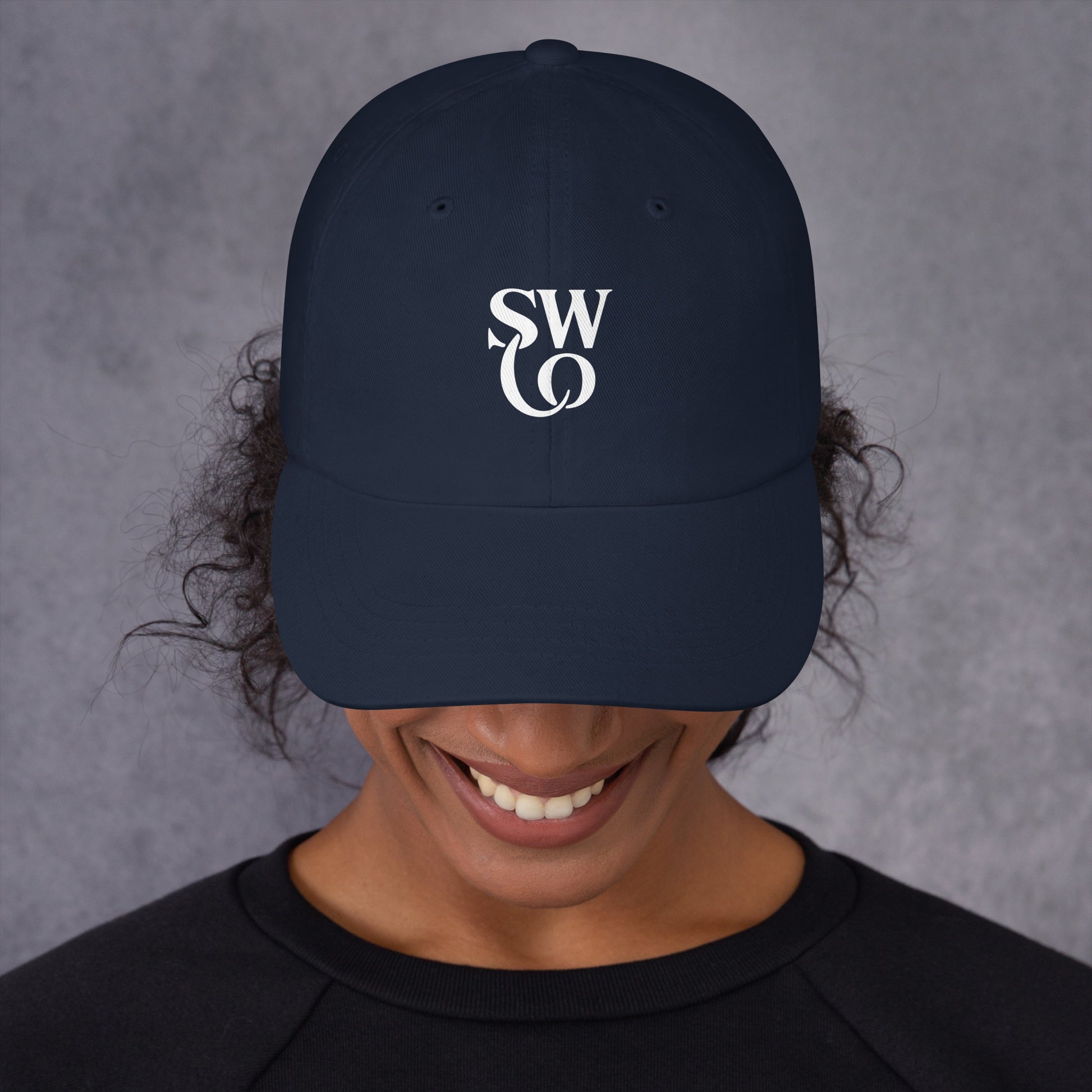 smiling person wearing navy dad hat with white SWO logo on front against gray background