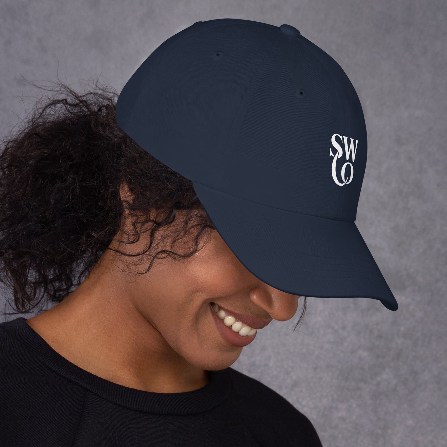 Smiling woman wearing a navy classic dad hat with white SWO logo on the side against gray background