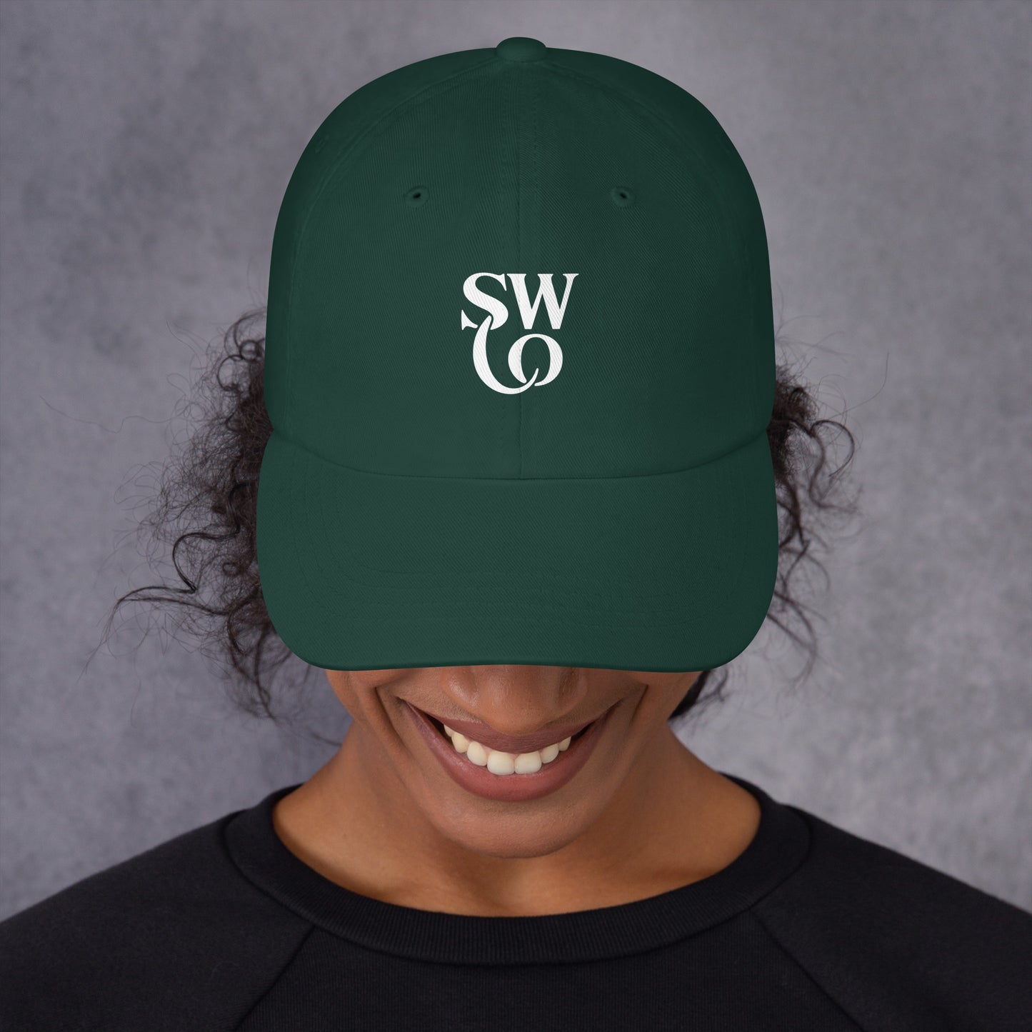 Smiling person wearing a spruce green dad hat with white stylish 'SWO' logo on front