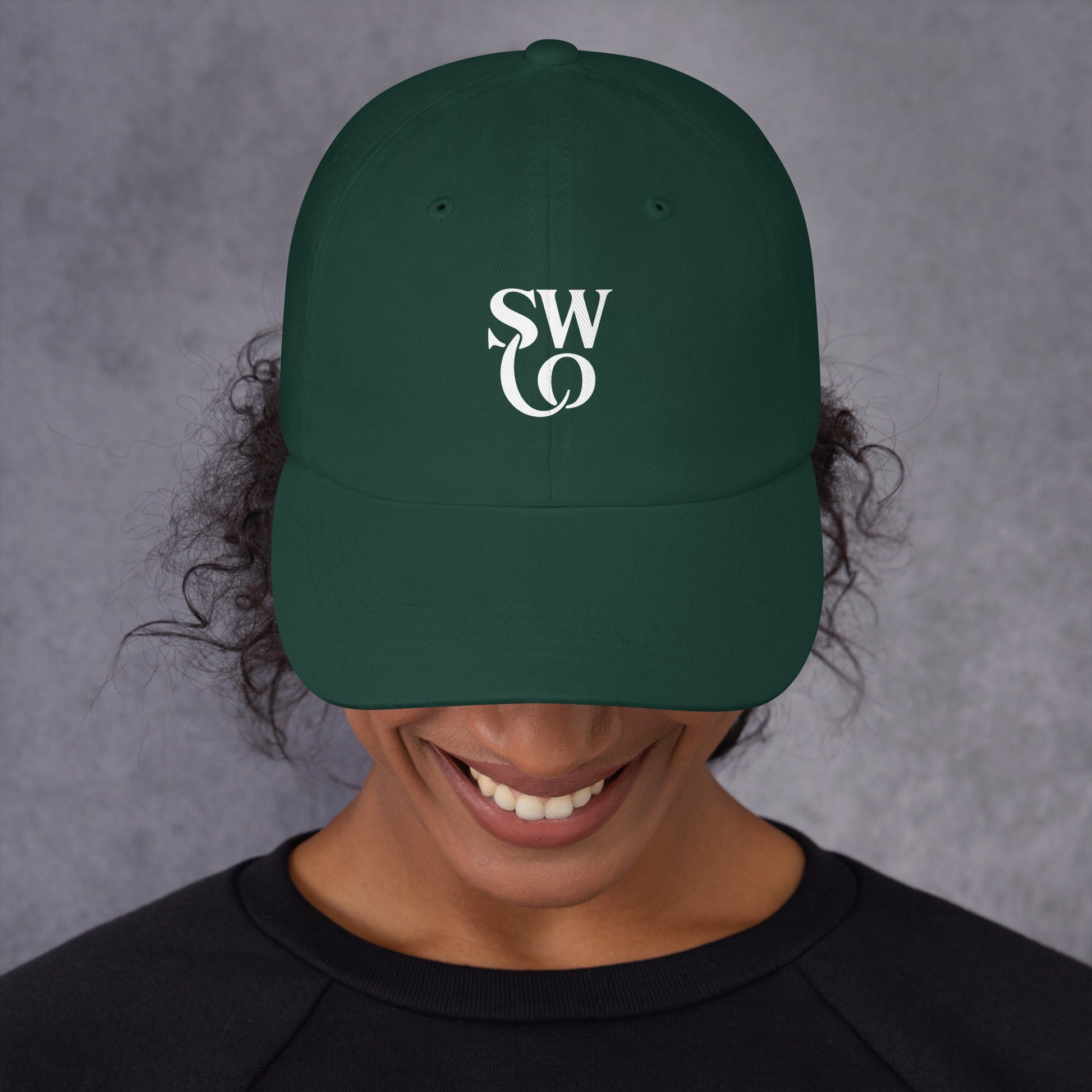 Smiling person wearing a spruce green dad hat with white stylish 'SWO' logo on front