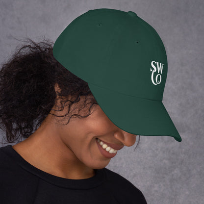 Smiling woman wearing a spruce green classic dad hat with white SWO logo on gray background