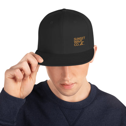 Man wearing black classic snapback hat with brown Sunset Wave Co. logo, navy sweatshirt