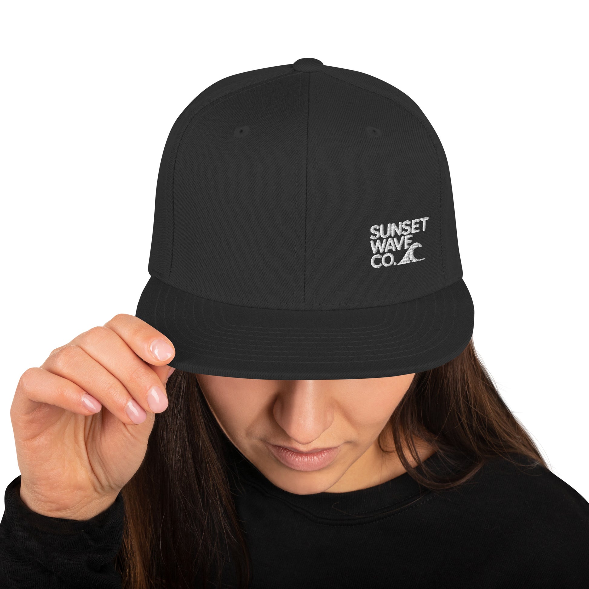Woman wearing a black snapback hat with white Sunset Wave Co. logo, black top, holding hat brim