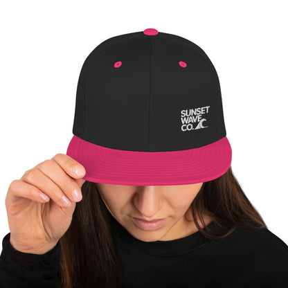 Black snapback hat with neon pink brim and details, white embroidered "Sunset Wave Co." logo, worn by woman
