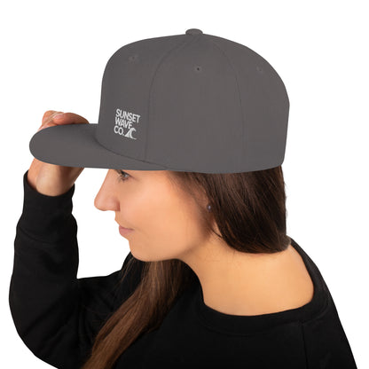 Woman wearing dark grey snapback cap with white Sunset Wave Co. logo, black sweatshirt