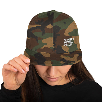 Woman wearing green camo snapback hat with white Sunset Wave Co. logo
