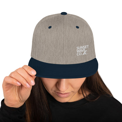 Heather grey and navy snapback hat with Sunset Wave Co. logo worn by woman holding brim