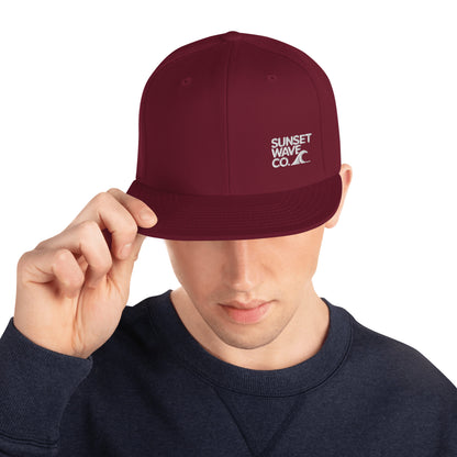 Maroon classic snapback hat with white Sunset Wave Co. logo worn by man in navy sweatshirt
