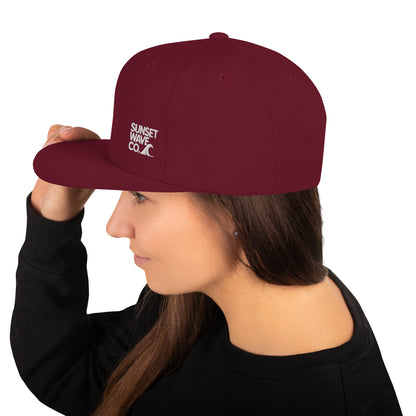 Woman wearing maroon classic snapback cap with white Sunset Wave Co. logo on side