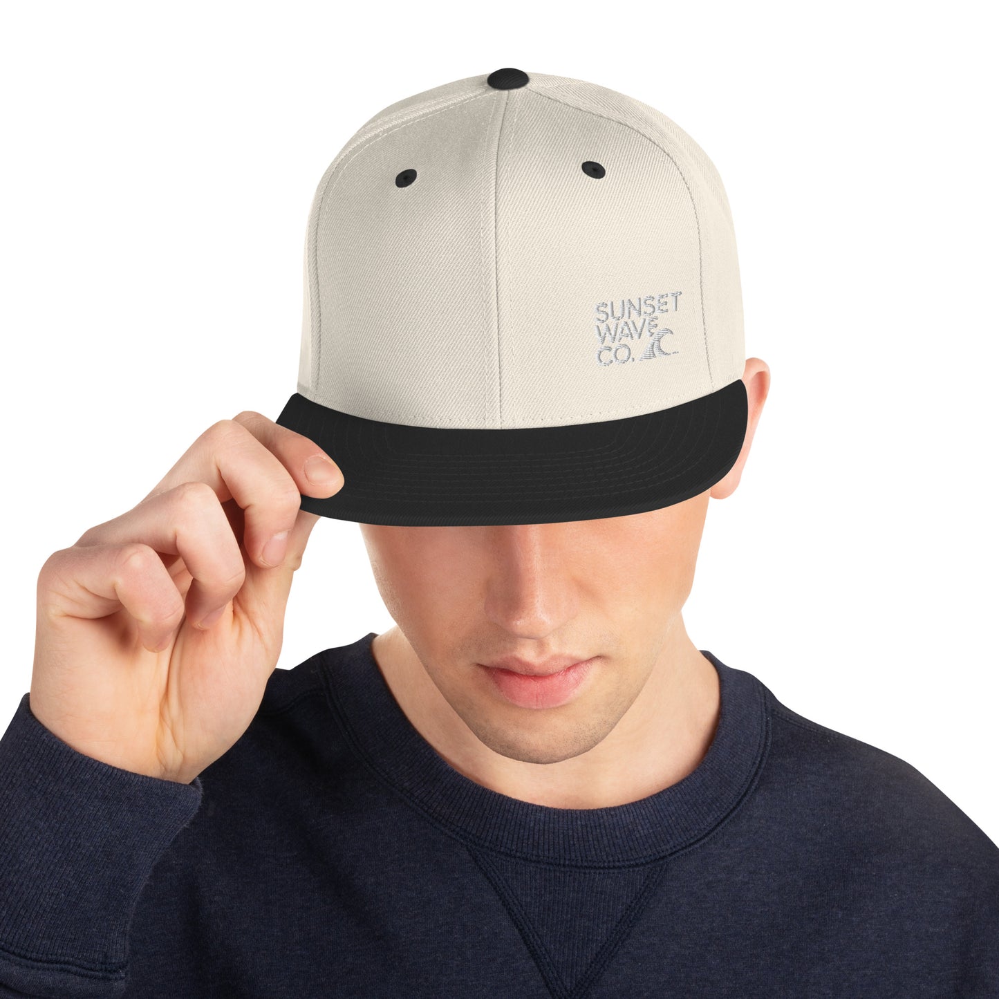 Man wearing beige and black classic snapback hat with Sunset Wave Co. logo, navy sweatshirt