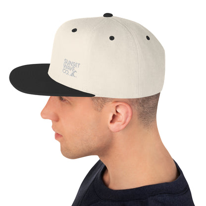 Side view of man wearing beige and black classic snapback hat with Sunset Wave Co. logo