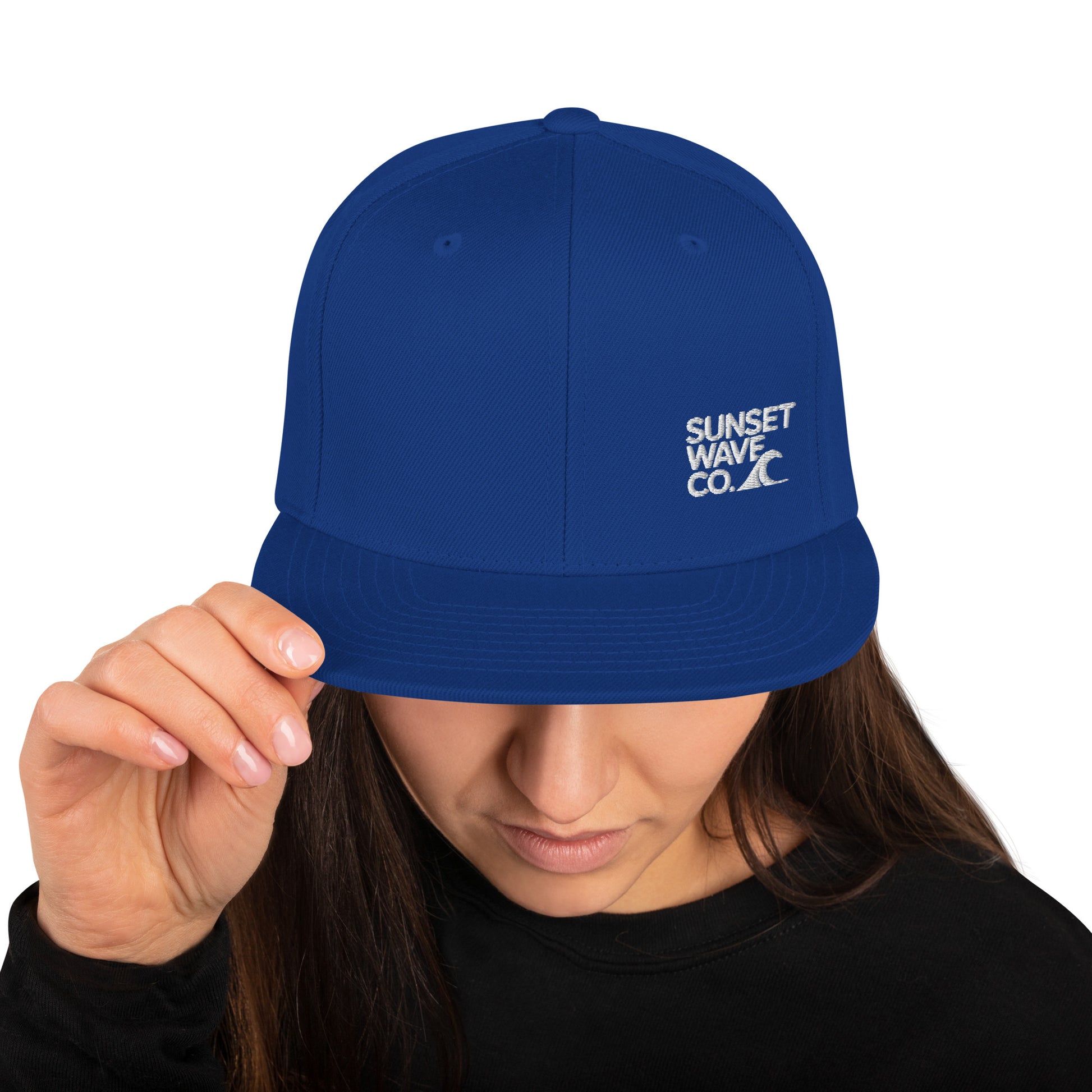 Woman wearing royal blue classic snapback hat with white Sunset Wave Co. logo, holding brim