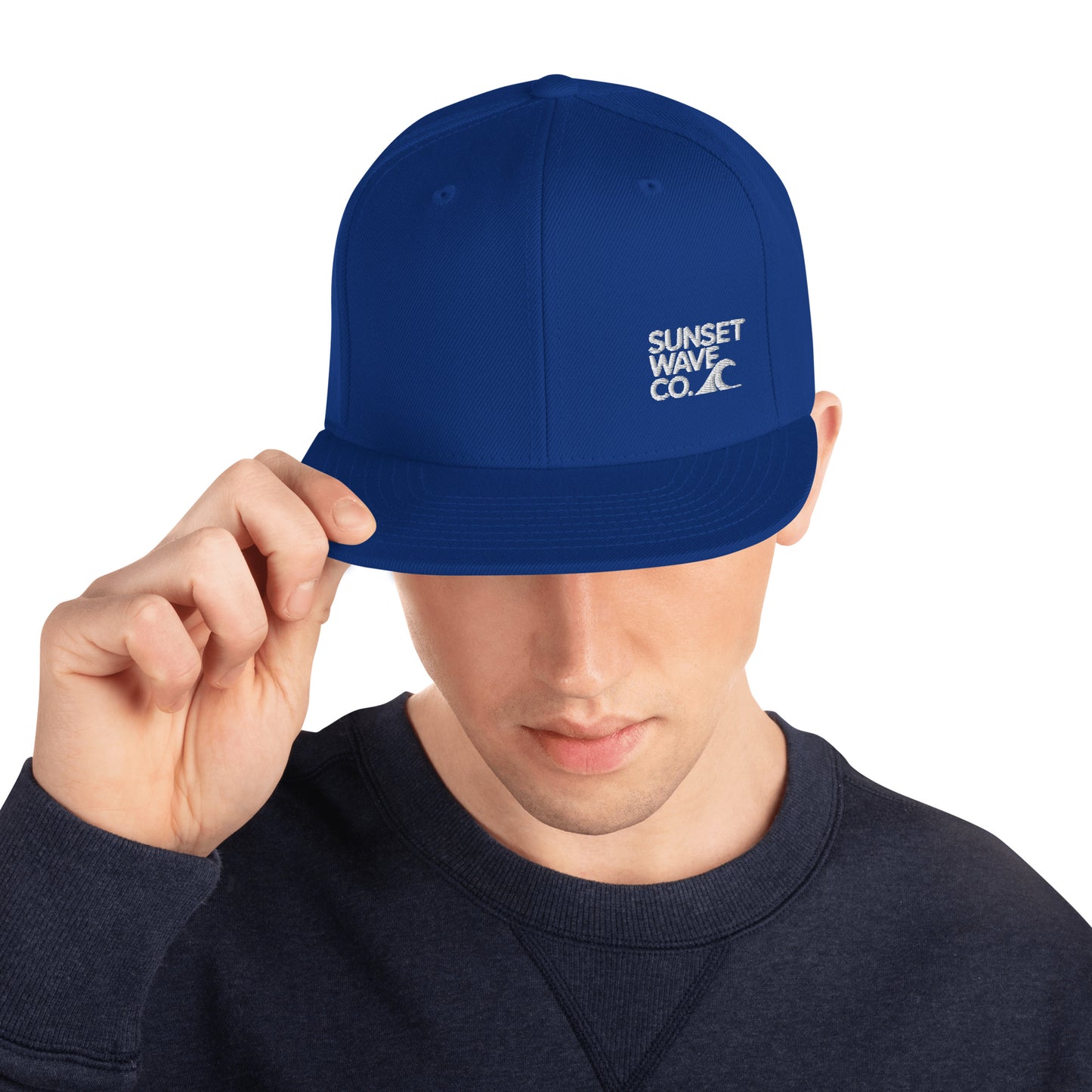 Man wearing royal blue classic snapback hat with white Sunset Wave Co. logo, adjusting brim