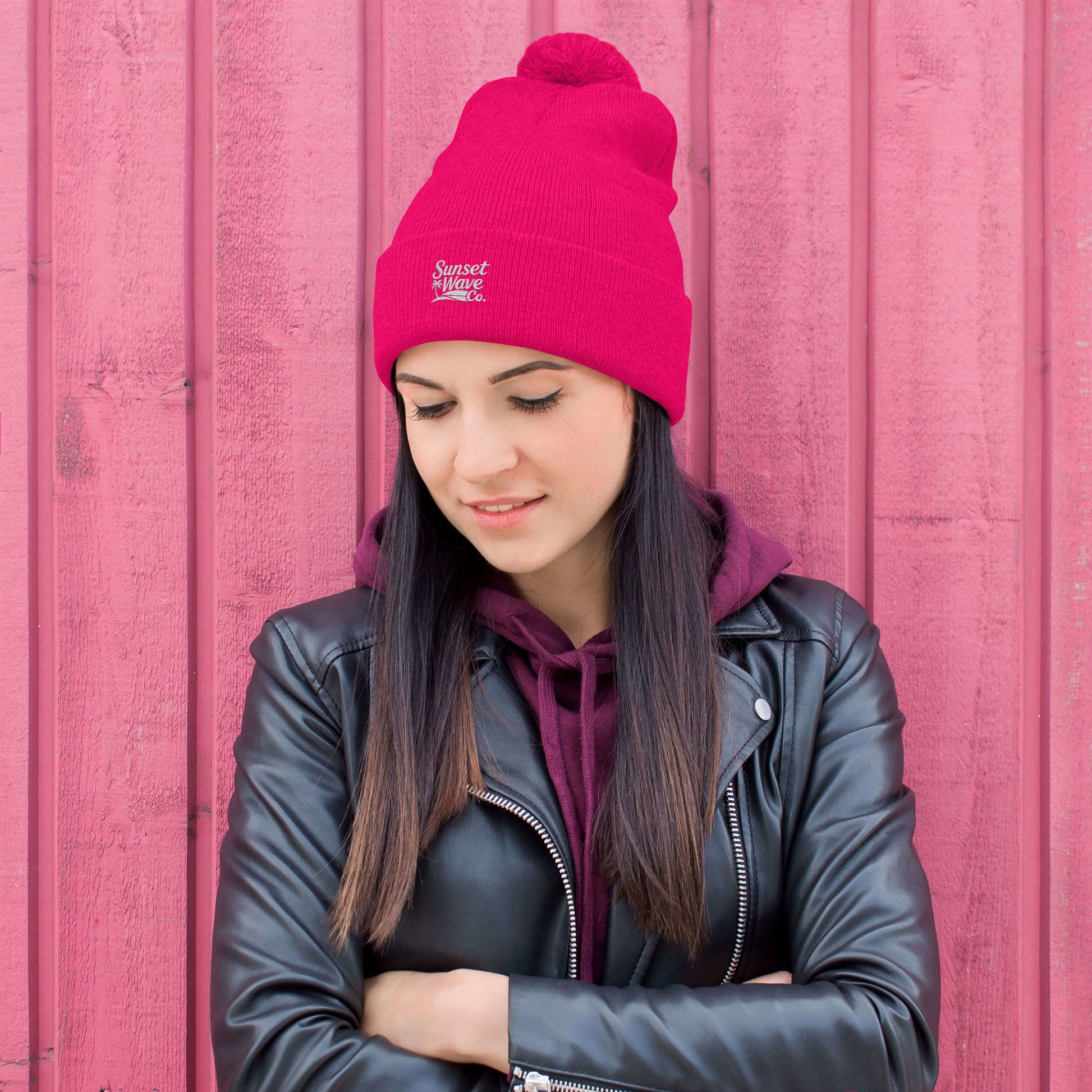 Young woman wearing neon pink pom-pom knit cap and black leather jacket against pink wooden wall