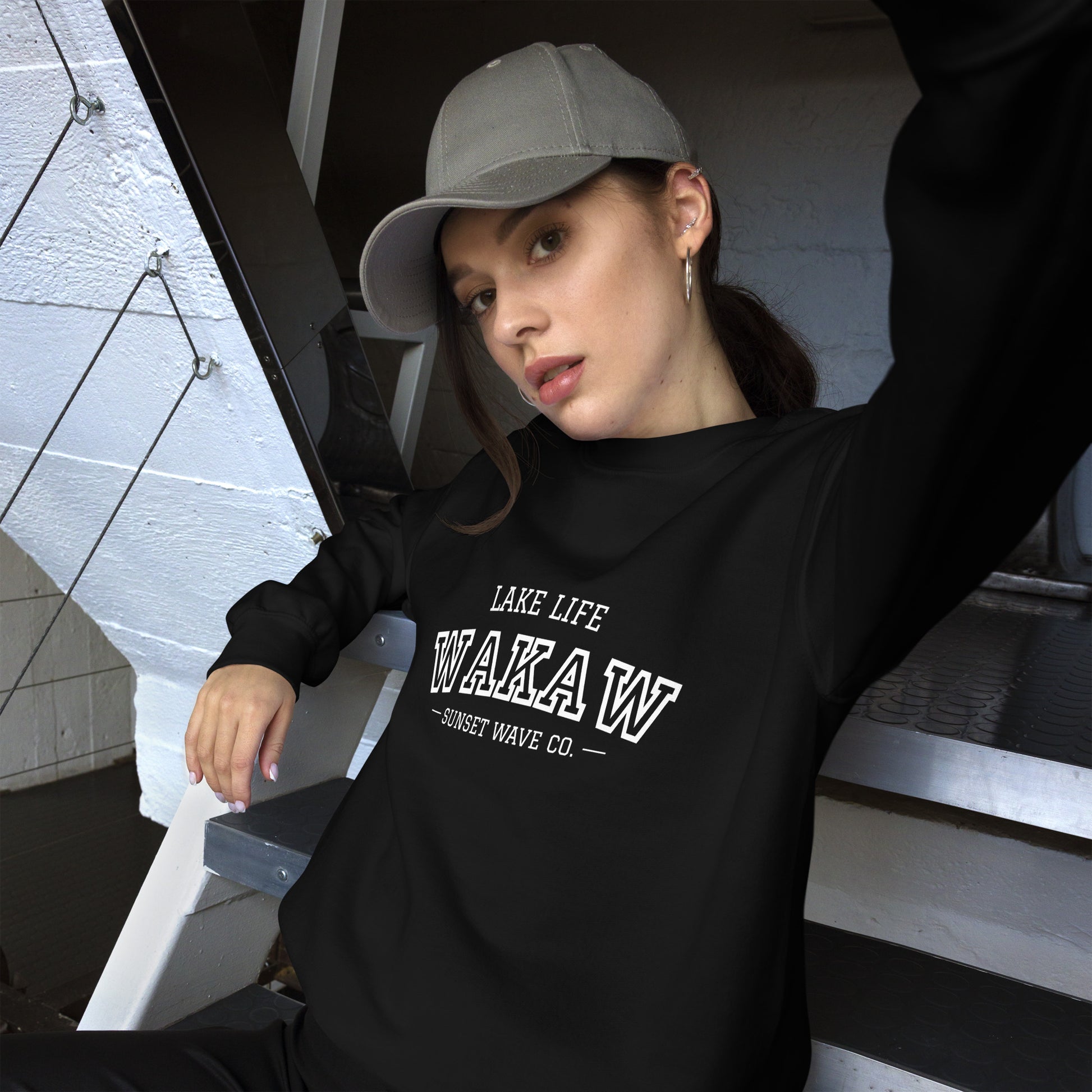 Woman wearing black crew neck sweatshirt with 'Lake Life WAKAW Sunset Wave Co.' text and gray cap sitting on metal stairs