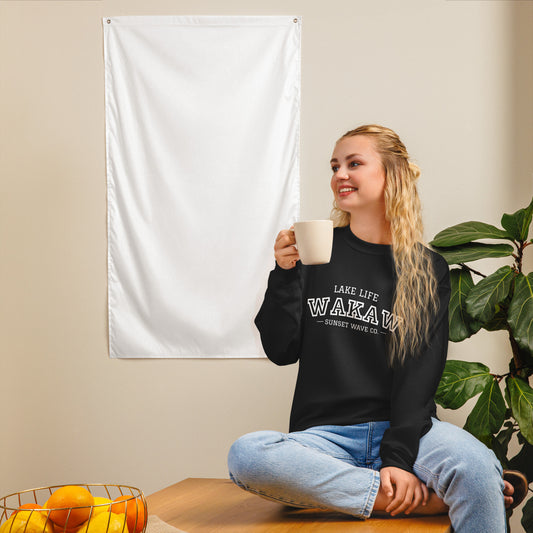 Smiling woman wearing black Lake Life WAKAW sweatshirt, holding white mug, sitting cross-legged by plant and fruit basket