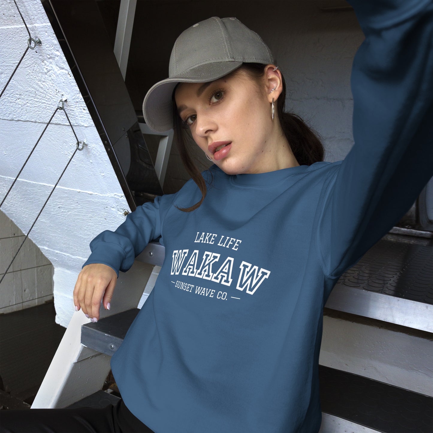 Woman wearing indigo blue unisex sweatshirt with white 'WAKA W' text, gray cap, sitting on metal stairs