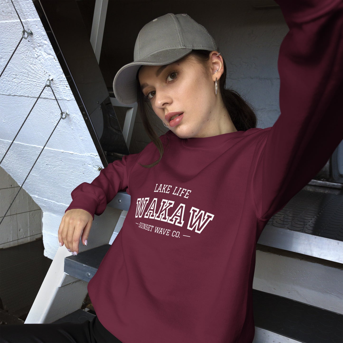 young woman wearing maroon unisex crew neck sweatshirt with Lake Life Waka W text and grey cap sitting on metal stairs