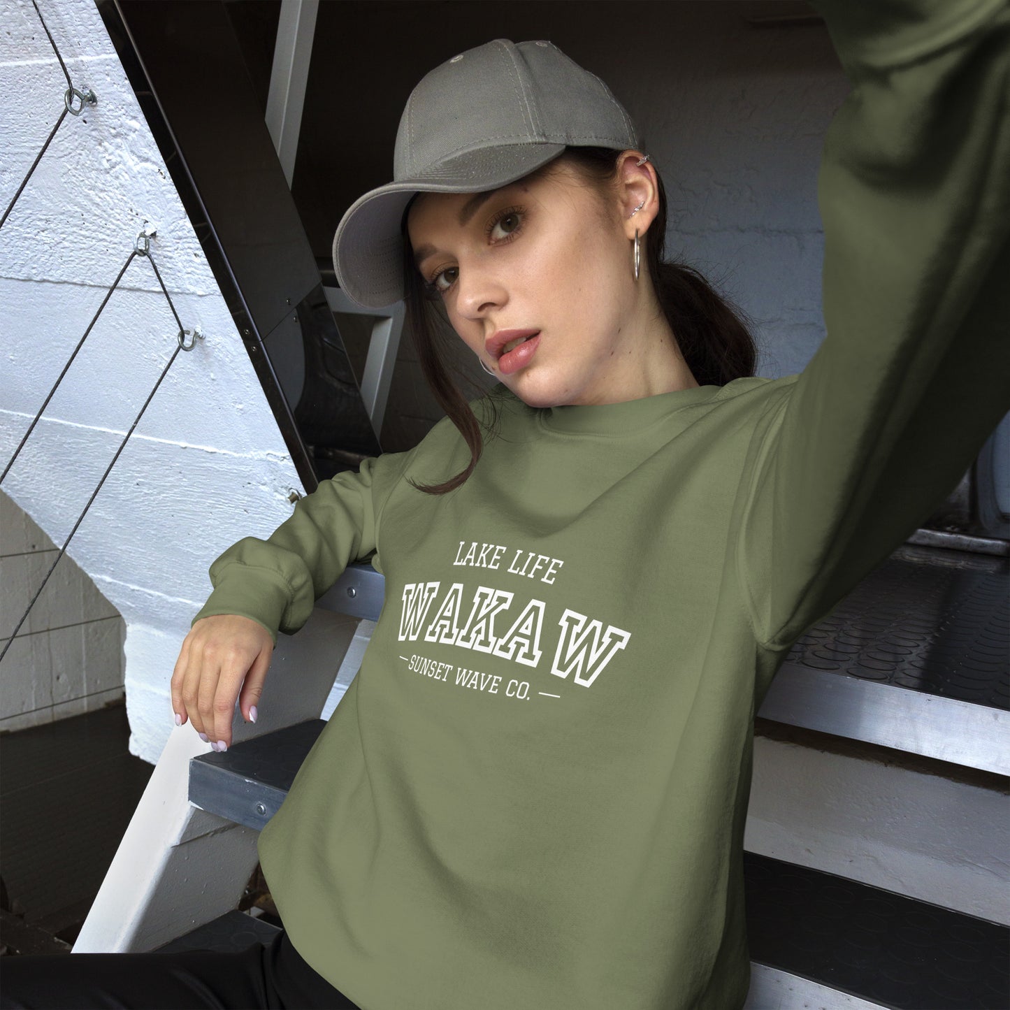 Young woman in military green crew neck sweatshirt with WAKAW text, posing on indoor metal stairs