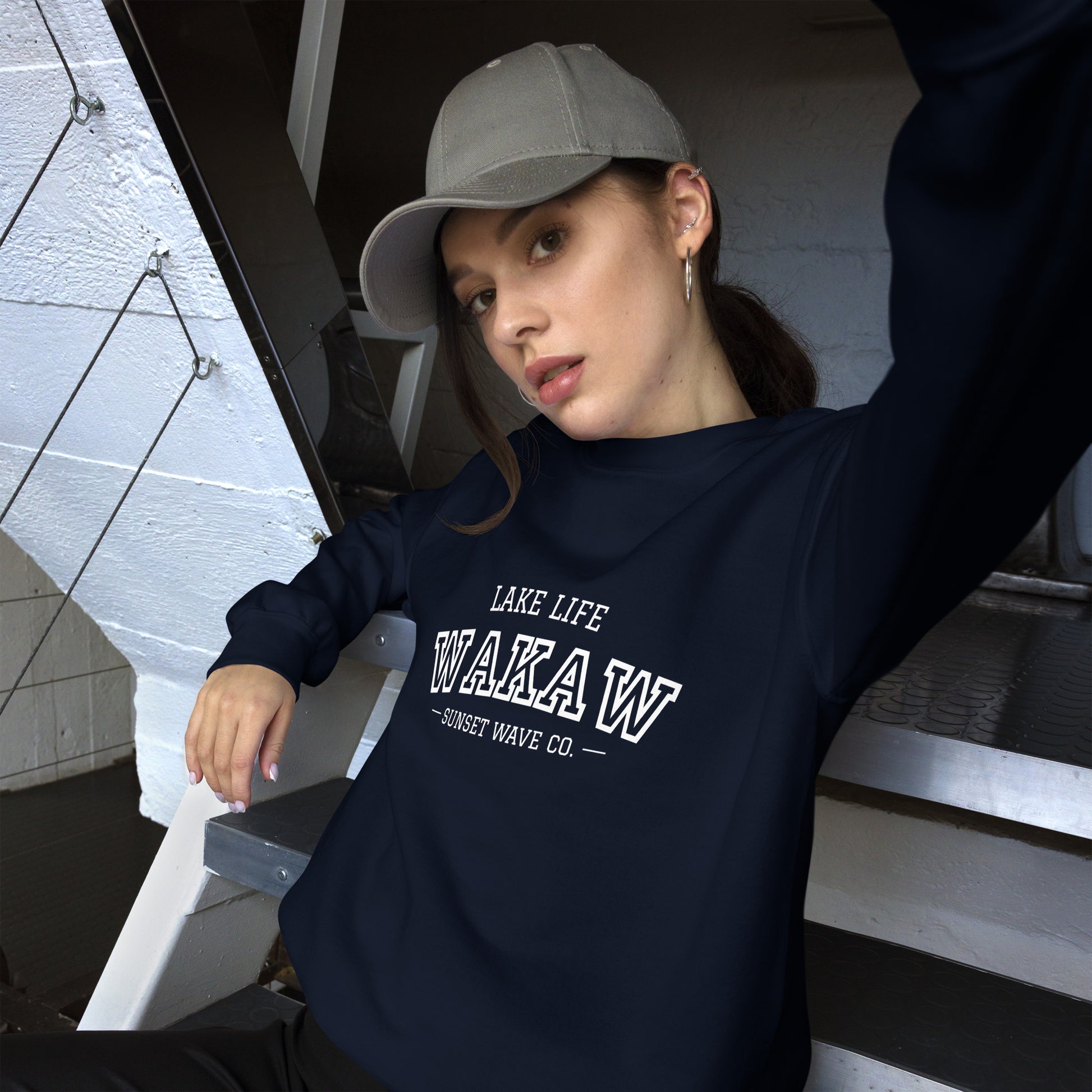 Young woman wearing navy sweatshirt with WAKA W lake life logo and gray baseball cap sitting on metal stairs