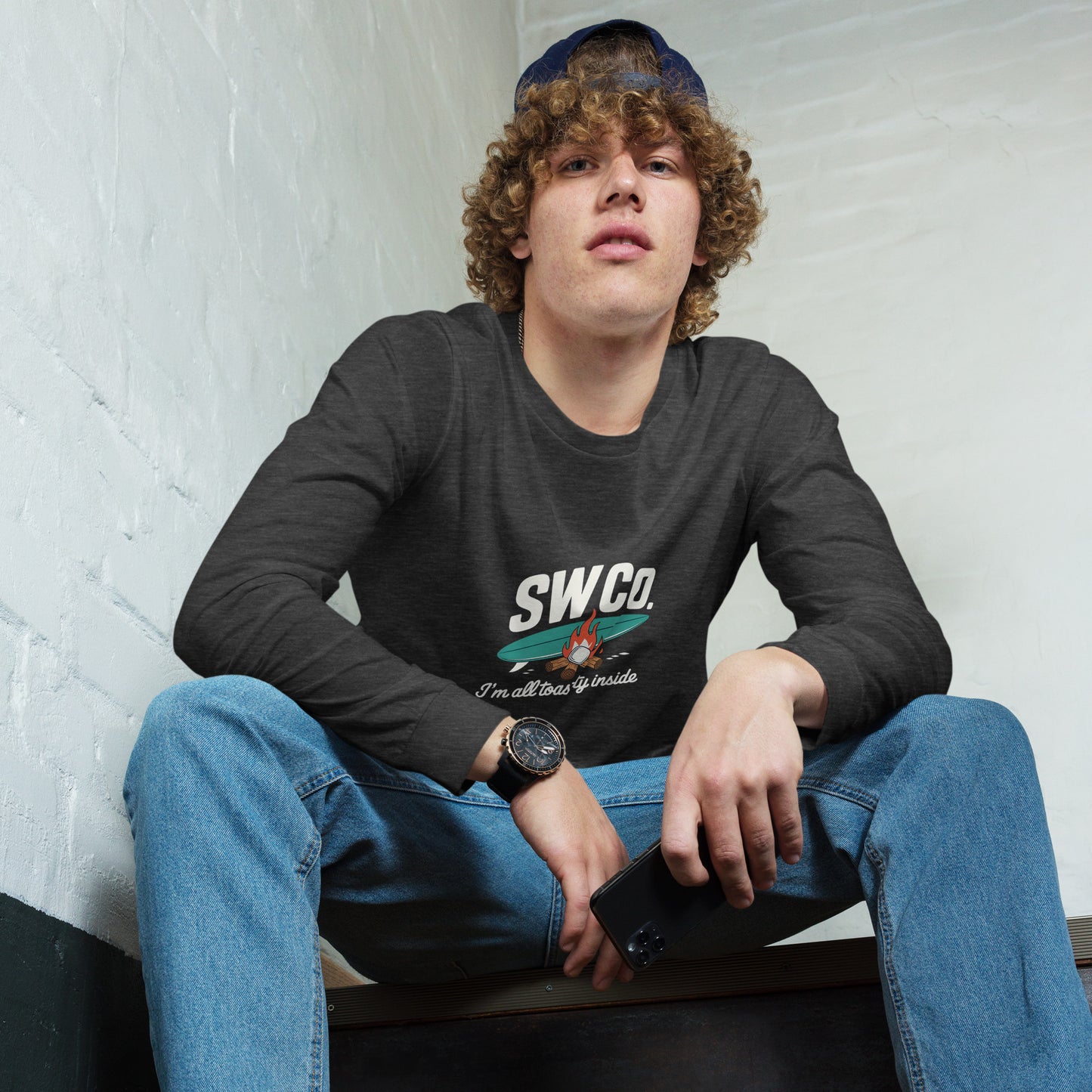 Young man wearing dark grey heather long sleeve tee with SWCo logo and graphic, blue jeans, black watch, holding black smartphone