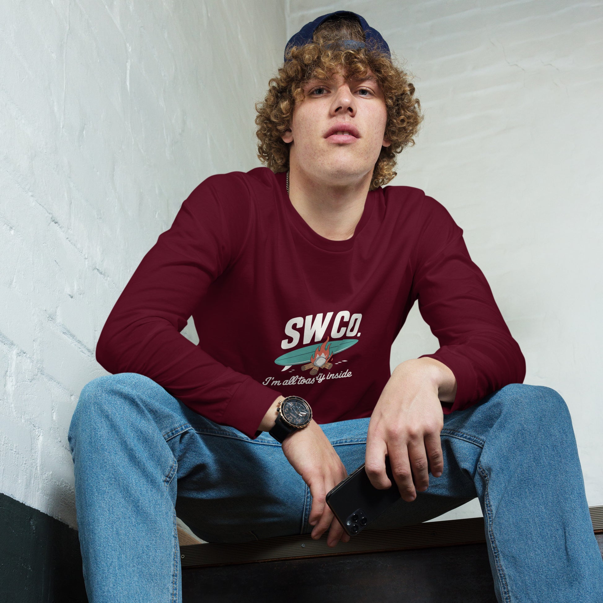 Young man wearing maroon SW Co. graphic long sleeve tee sitting on stairs with blue jeans and cap