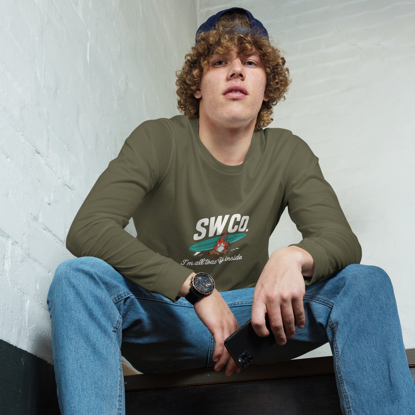 Young man wearing military green long sleeve tee with SWCO logo and campfire graphic, blue jeans, sitting indoors