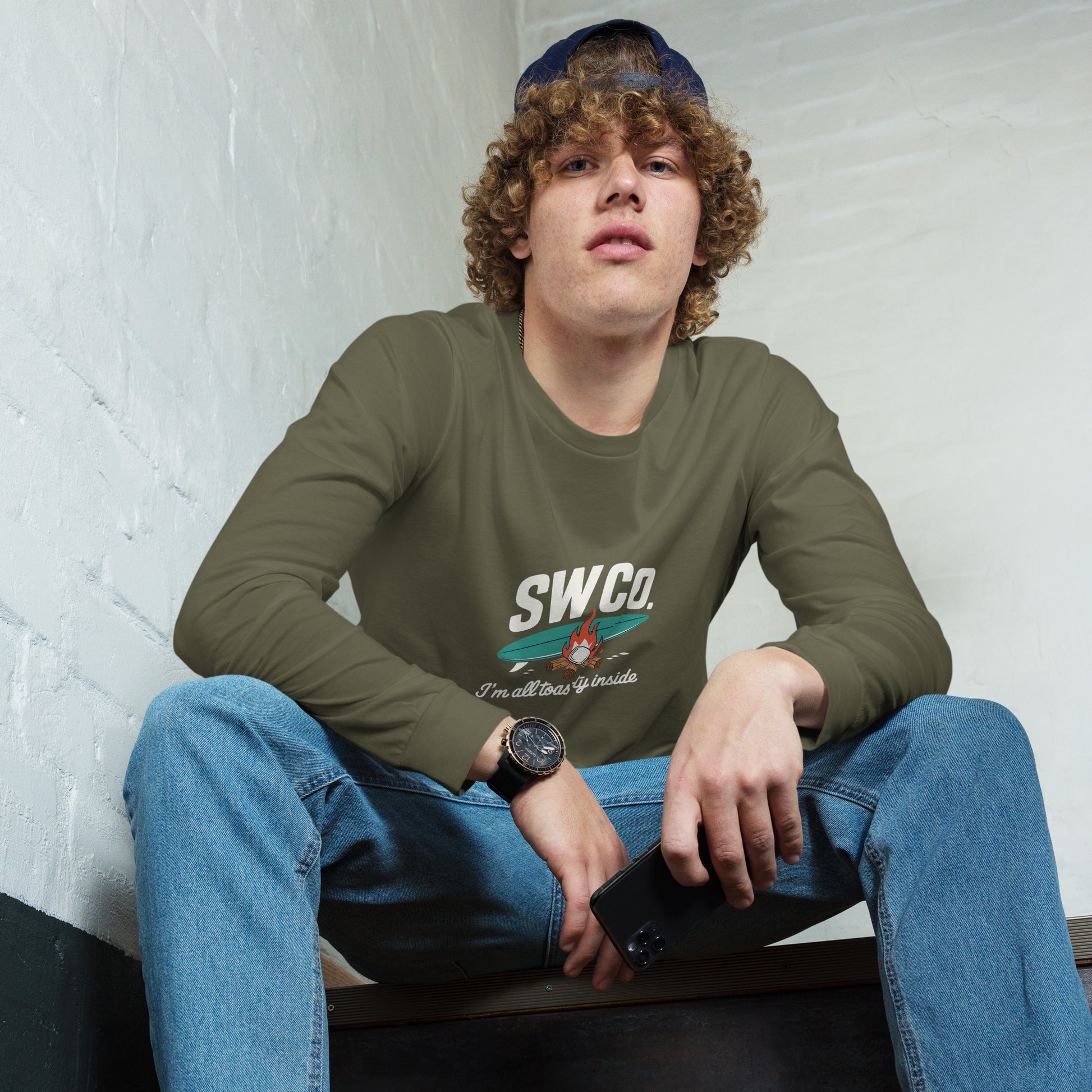 Young man wearing military green long sleeve tee with SWCO logo and campfire graphic, blue jeans, sitting indoors