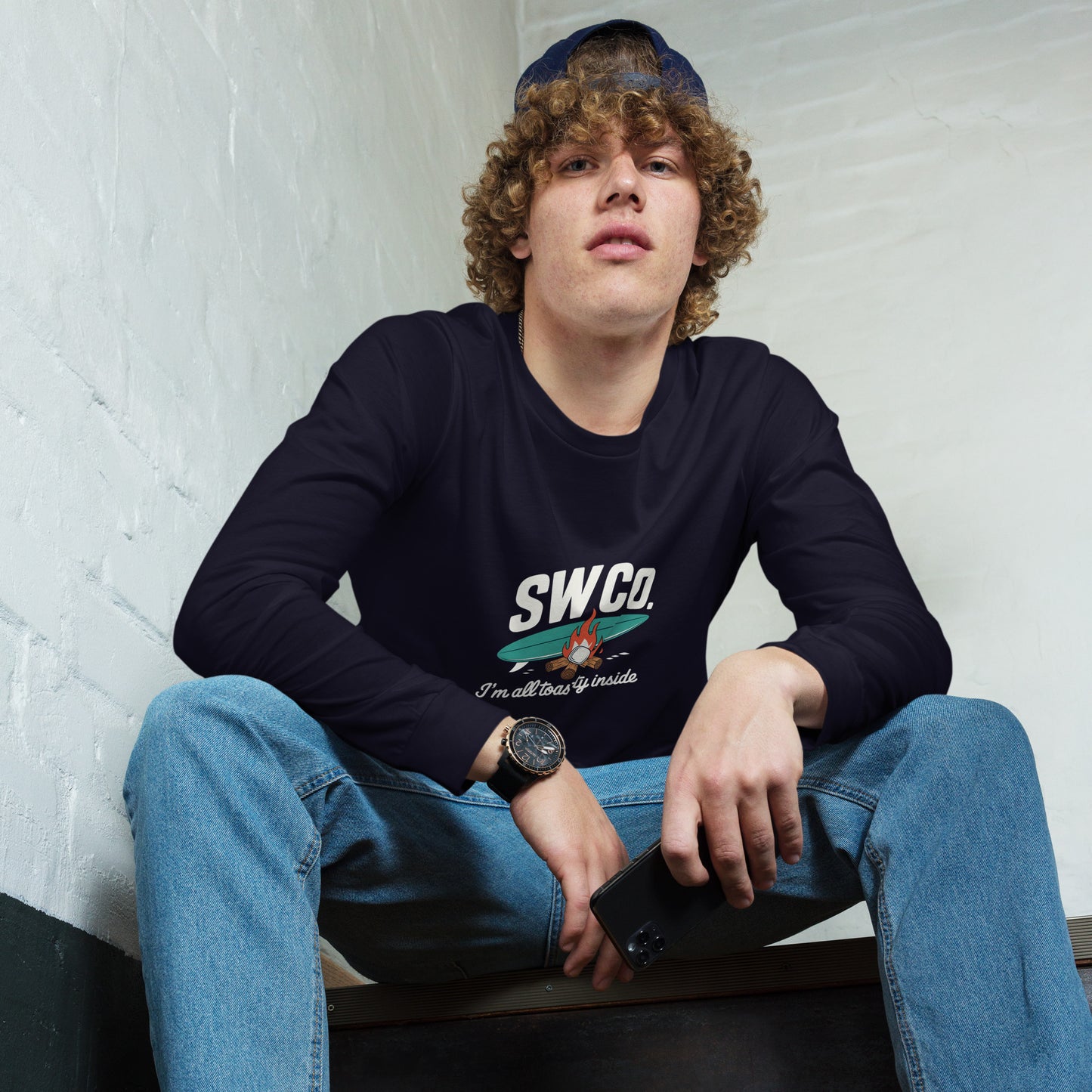 Young man wearing navy unisex long sleeve tee with SW Co. campfire logo, sitting indoors