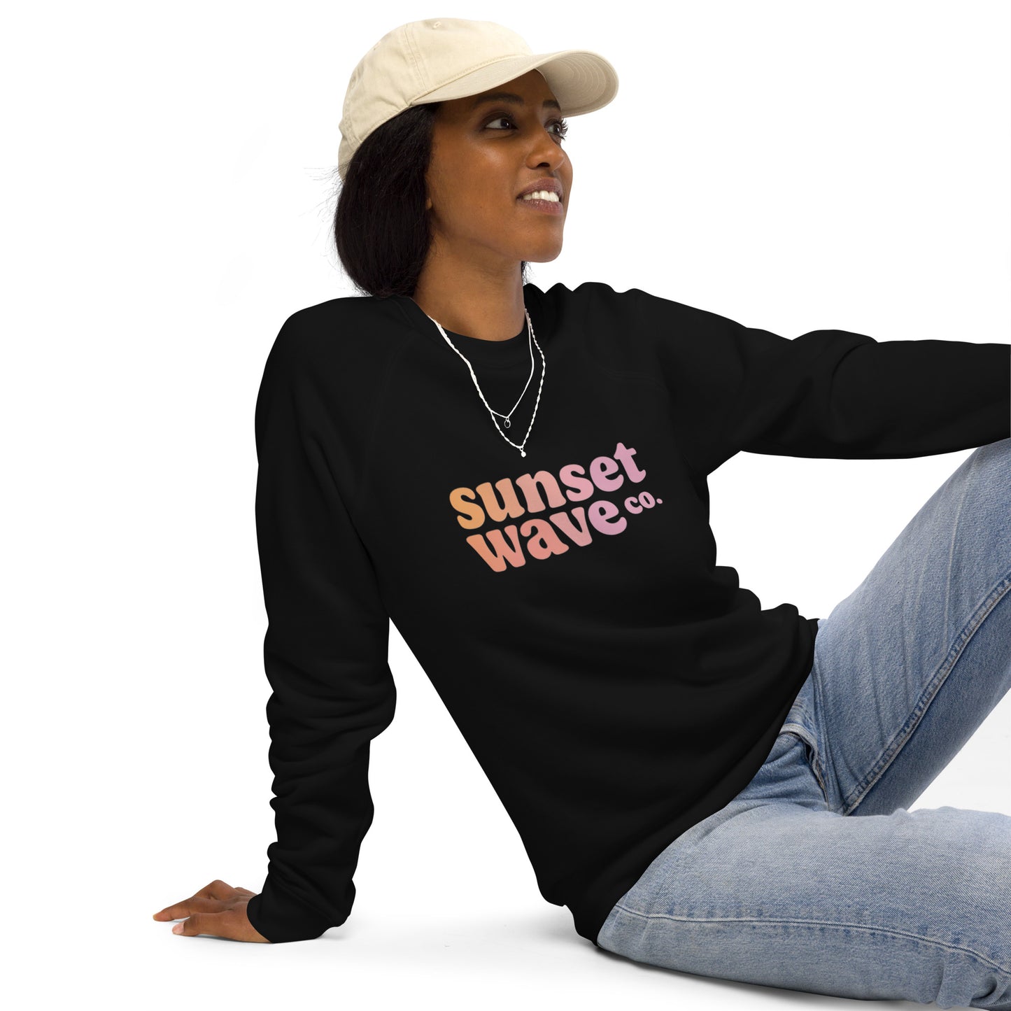 Woman wearing black unisex organic raglan sweatshirt with Sunset Wave Co. logo, cream cap, and light blue jeans