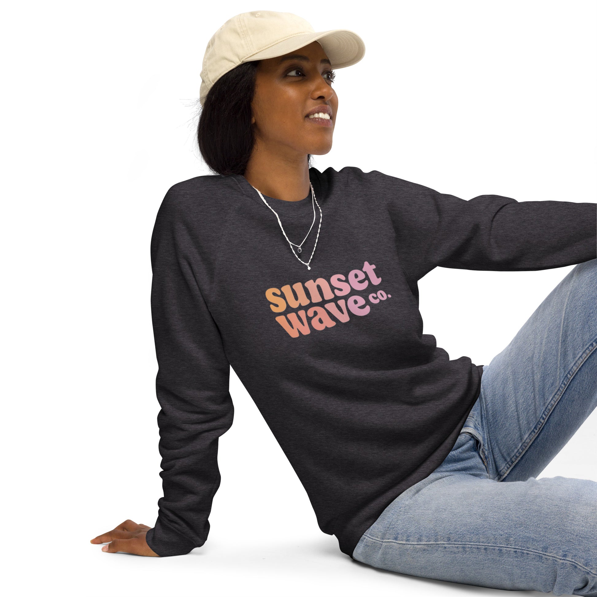 woman wearing charcoal melange sweatshirt with sunset wave co. print sitting casually in jeans and beige cap