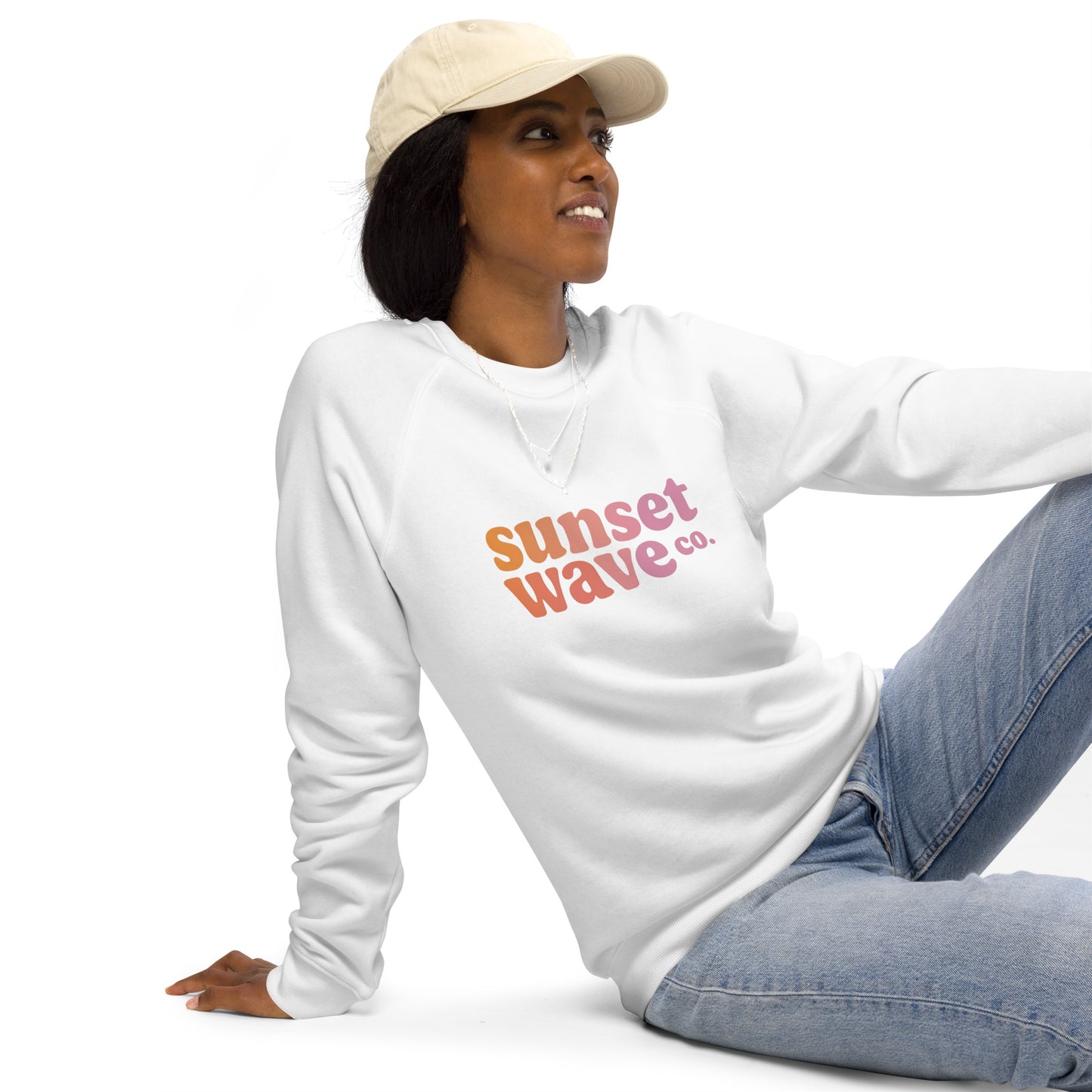 Woman wearing white unisex organic raglan sweatshirt with Sunset Wave Co. logo sitting on floor in casual jeans and beige cap