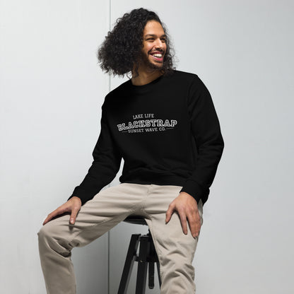 Smiling man wearing black unisex sweatshirt with 'Lake Life Blackstrap Sunset Wave Co.' text, beige pants, sitting on stool