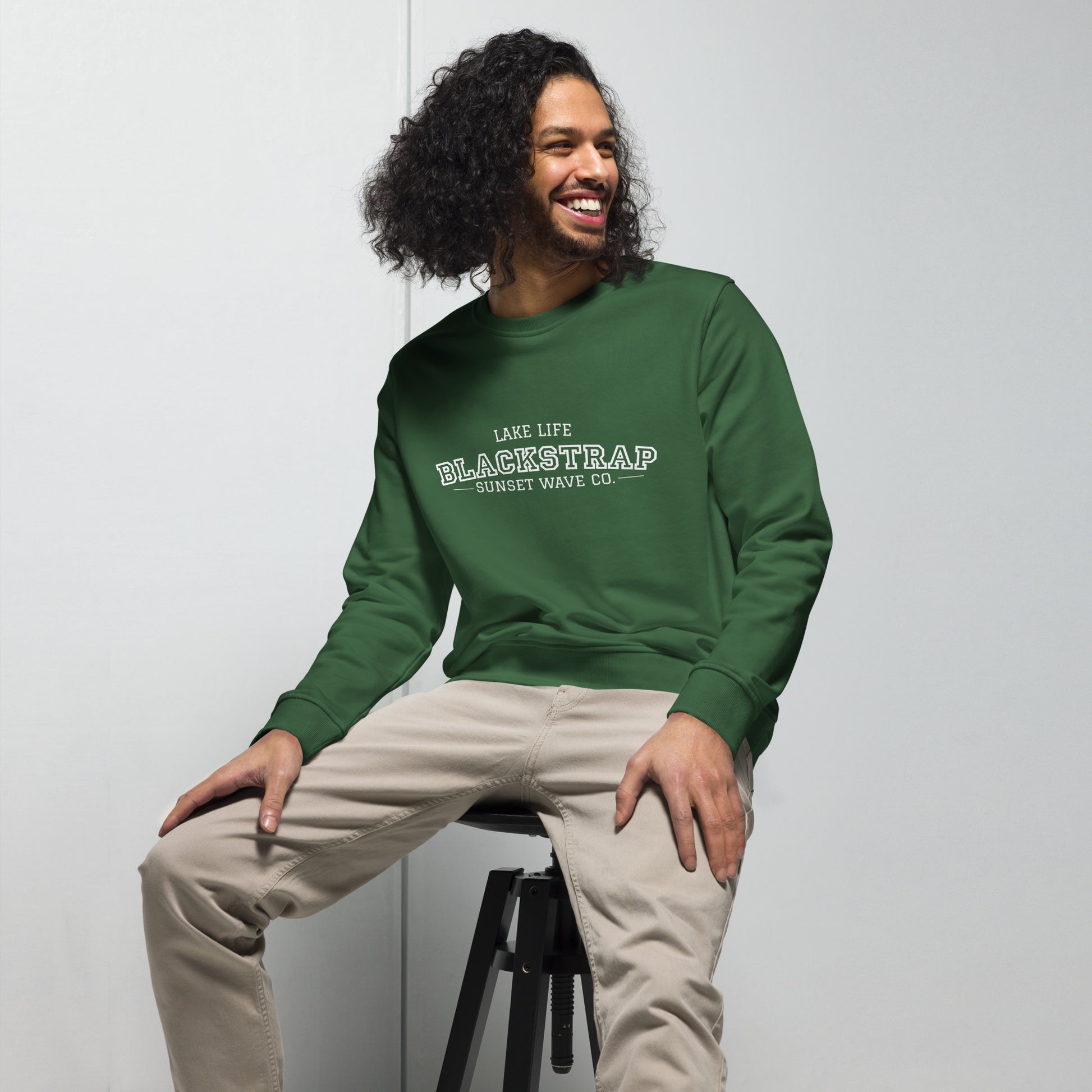 Man wearing unisex organic bottle green sweatshirt with white Blackstrap logo sitting on stool