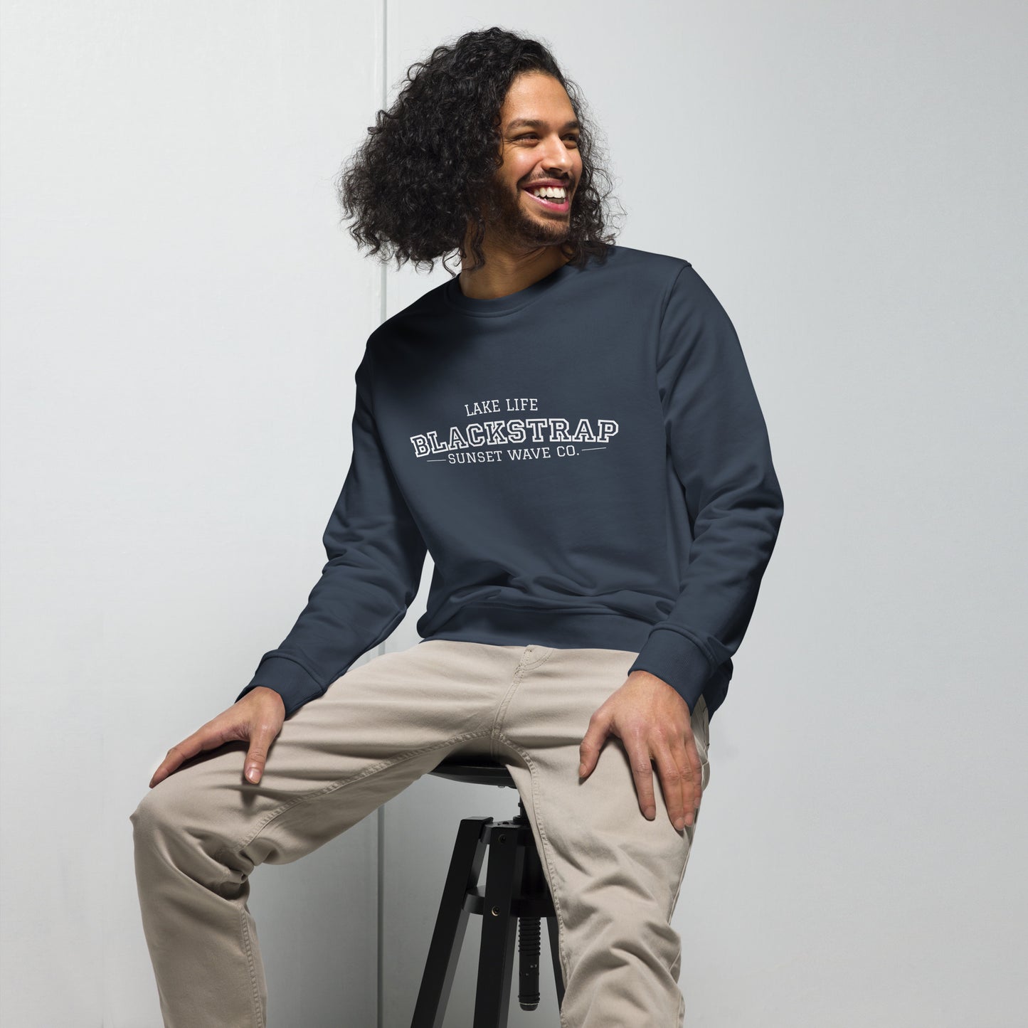 Smiling man with curly hair wearing a French navy organic sweatshirt with 'Blackstrap' logo sitting on a stool