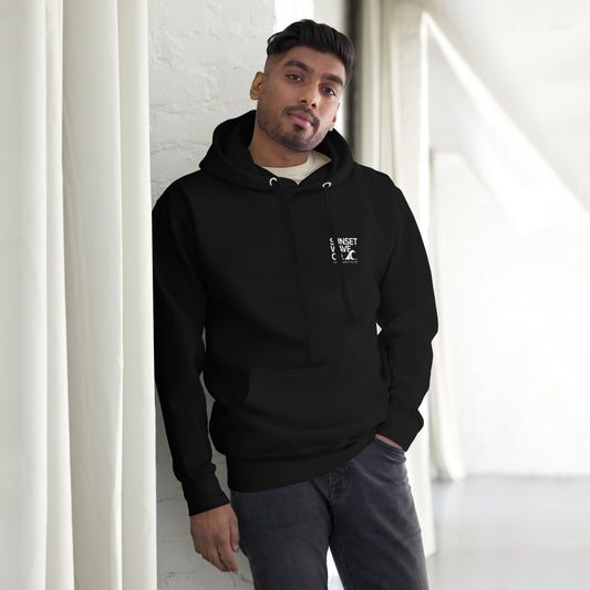 Man wearing black unisex premium hoodie with small white Sunset Wave Co logo, standing indoors
