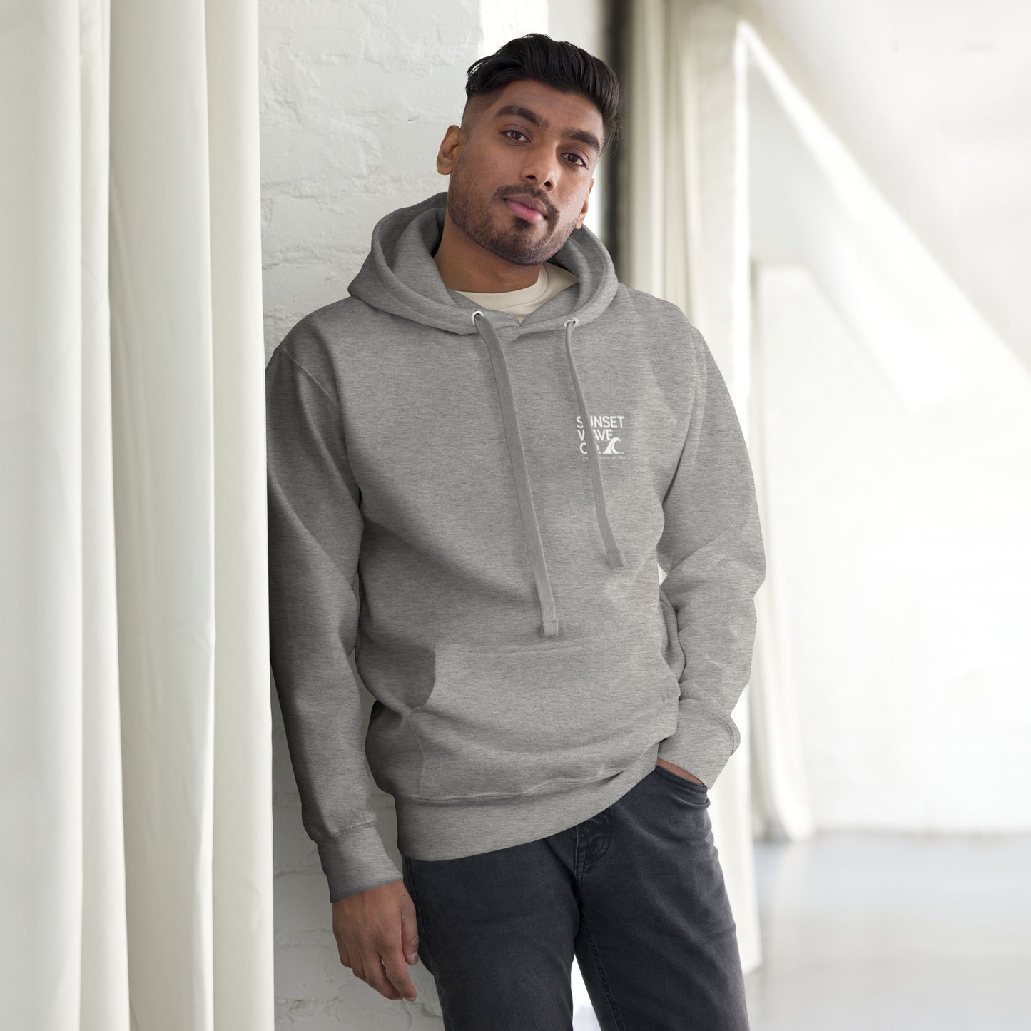 man wearing unisex premium grey hoodie with white text print, posing indoors by white curtains