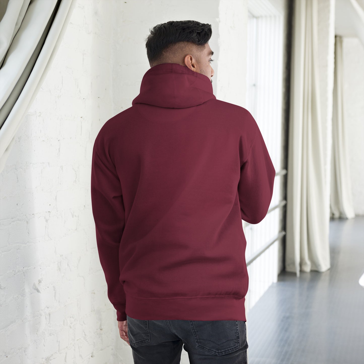 Back view of man wearing maroon unisex premium hoodie indoors with white brick wall and curtains