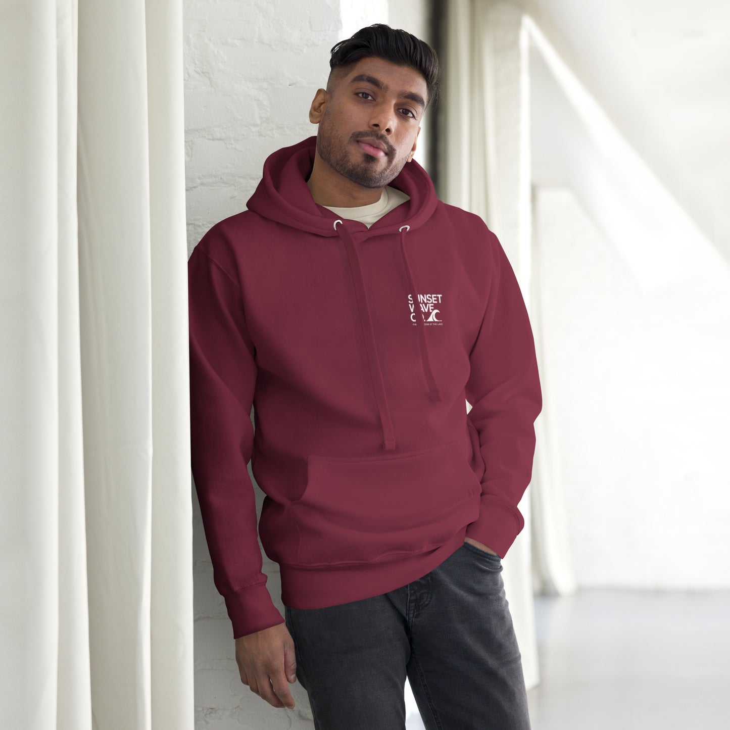 Man wearing maroon unisex premium hoodie with white sunset wave logo in minimalist bright room